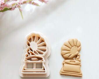 May include: A white plastic cookie cutter and a brown cookie in the shape of a vintage gramophone.