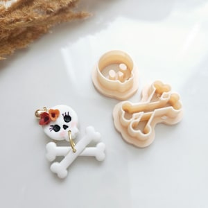 May include: White sugar cookie cutters in the shape of a skull and crossbones. The skull cutter has two small holes for eyes and a larger hole for the mouth. The crossbones cutter has two bones that are crossed at the center.
