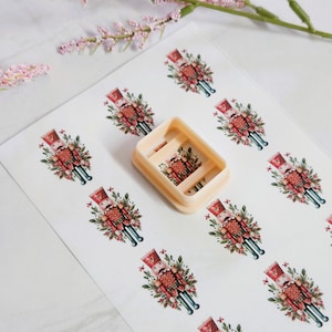 May include: A beige rectangular cookie cutter sits on a sheet of paper with repeating images of a Christmas nutcracker soldier. The nutcracker is dressed in a red uniform with gold accents and is surrounded by greenery and flowers. The background is white.