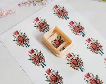 May include: A beige rectangular cookie cutter sits on a sheet of paper with repeating images of a Christmas nutcracker soldier. The nutcracker is dressed in a red uniform with gold accents and is surrounded by greenery and flowers. The background is white.