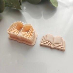 May include: A set of four plastic cookie cutters shaped like an open book. The cutters are layered, with the largest cutter on the bottom and the smallest cutter on top. The cutters are a light peach color.