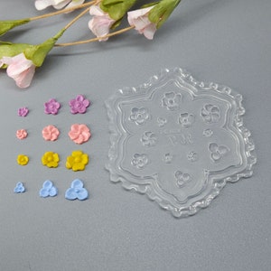 May include: A clear, flower-shaped silicone mold with various flower-shaped embellishments in pink, purple, yellow, and blue. The mold has a decorative border and several flower impressions. A sprig of pink flowers is in the background.