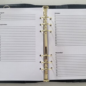 Puede incluir: Una agenda encuadernada en cuero negro con anillos dorados y un bolígrafo dorado. La agenda tiene páginas para agosto, septiembre, octubre y noviembre. La palabra "Hoy" está escrita en la parte superior de la página.