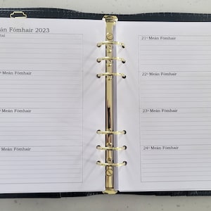 May include: Open planner with gold accents, featuring lined pages for notes and a calendar for Meán Fómhair 2023. The planner is dark blue with a textured finish and a gold zipper. A gold pen is attached to the side.