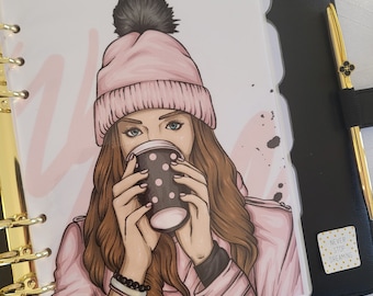 May include: A black planner with gold accents, a gold heart-shaped paper clip, and a gold pen. The planner features a decorative insert with a woman wearing a pink hat, drinking from a black polka-dot cup. The words "Never Stop Dreaming" are on a sticker.