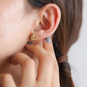 May include: Gold pumpkin-shaped earrings with a jack-o'-lantern face cutout. The earrings are worn in a person's ear.