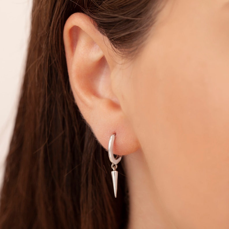 Puede incluir: Pendientes de aro de plata con una espiga de plata colgante. Los pendientes se llevan en la oreja de una persona.