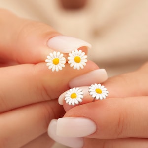 May include: A pair of silver stud earrings with a white daisy design and a yellow center. The earrings are shown on a person's hand.