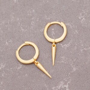Puede incluir: Un par de pendientes de aro de oro con un colgante puntiagudo y afilado. Los pendientes son simples y elegantes, con un dise&ntilde;o moderno.