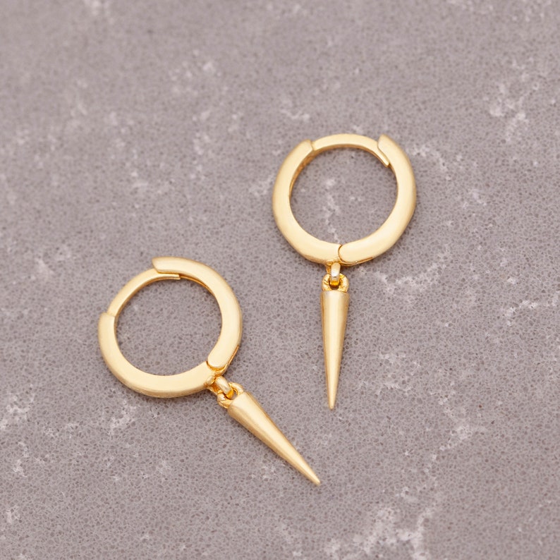 Puede incluir: Un par de pendientes de aro de oro con un colgante puntiagudo y afilado. Los pendientes son simples y elegantes, con un dise&ntilde;o moderno.