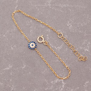 May include: A gold chain bracelet featuring a blue and white evil eye charm. The charm is set with small clear stones.