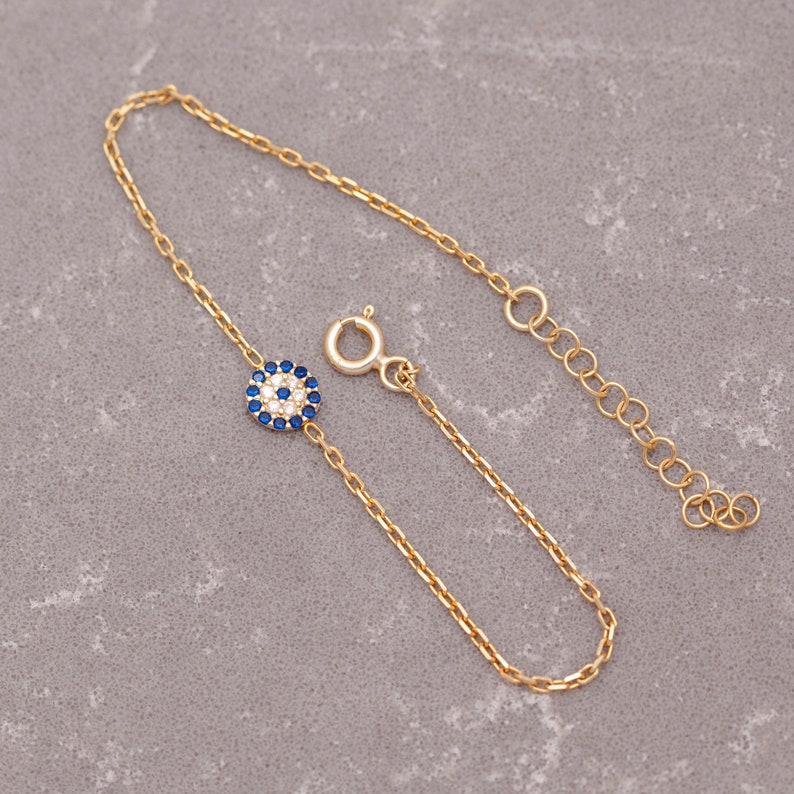 May include: A gold chain bracelet featuring a blue and white evil eye charm. The charm is set with small clear stones.