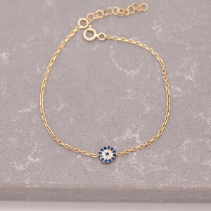May include: A gold chain bracelet featuring a small blue and white evil eye charm. The charm is surrounded by tiny white stones.