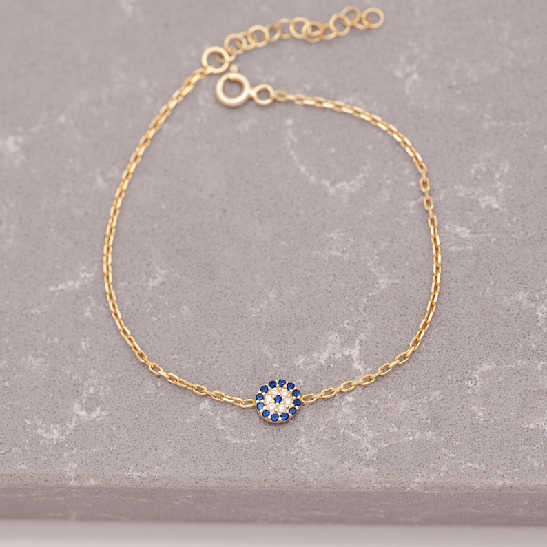 May include: A gold chain bracelet featuring a small blue and white evil eye charm. The charm is surrounded by tiny white stones.