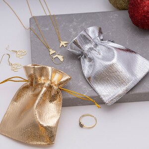 K&ouml;nnte beinhalten: Zwei goldene Schmuckbeutel, einer silber, einer gold, mit einer goldenen Halskette mit einem Schneemann- und Vogelanh&auml;nger, einem Paar goldenen Ohrringen und einem goldenen Ring mit einem schwarzen Stein.