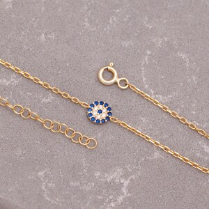 May include: A gold chain bracelet featuring a small blue and white evil eye charm. The charm is surrounded by tiny white stones.