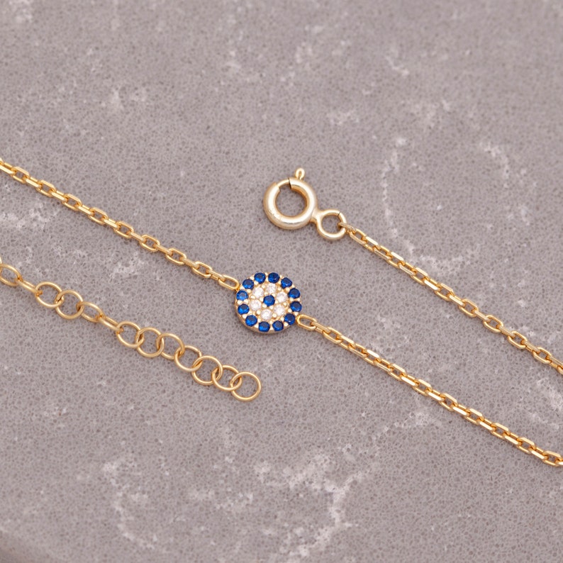 May include: A gold chain bracelet featuring a small blue and white evil eye charm. The charm is surrounded by tiny white stones.