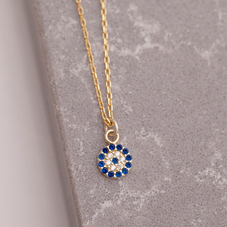 K&ouml;nnte beinhalten: Goldkette mit einem kleinen blauen und wei&szlig;en b&ouml;sen Blick Anh&auml;nger. Der Anh&auml;nger ist aus Gold und hat einen Kreis aus blauen Steinen um eine wei&szlig;e Mitte.