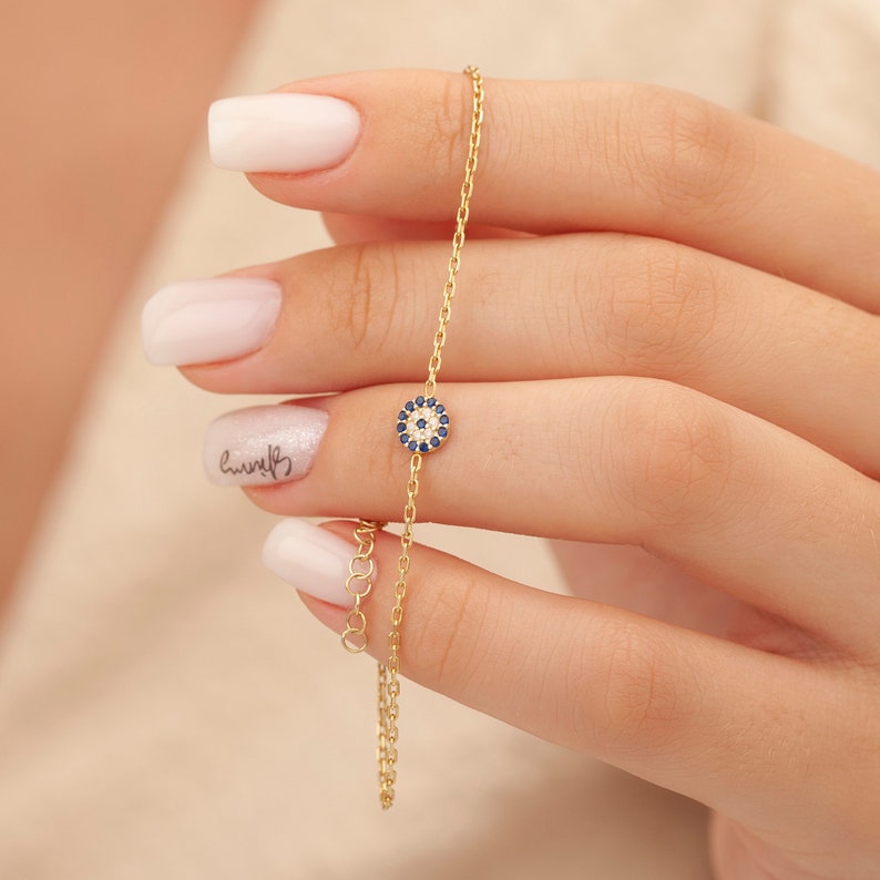 May include: A delicate gold chain bracelet featuring a small blue and white evil eye charm. The charm is surrounded by tiny white stones.