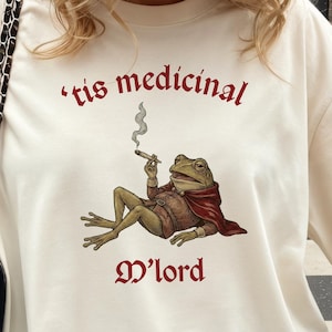 Może przedstawiać: Kremowa koszulka oversize z ilustracją żaby palącej papierosa, z bordowym napisem "'tis medicinal M'lord". Str&oacute;j uzupełnia beżowa czapka z daszkiem i czarna pikowana torba na ramię.