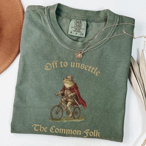 Könnte beinhalten: Ein olivgrünes Kurzarm-T-Shirt mit einer Grafik eines Frosches, der eine Krone und einen roten Umhang trägt und Fahrrad fährt. Der Text "Off to unsettle" steht über dem Frosch und "The Common Folk" darunter in einer Serifenschrift.