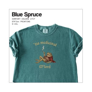 Może przedstawiać: Niebieska koszulka Blue Spruce w kolorze turkusowym. Koszulka przedstawia kresk&oacute;wkową żabę w pelerynie i palącą papierosa, z napisem "'tis medicinal M'lord". Koszulka jest Comfort Colors 1717 i dostępna w rozmiarach S-4XL.