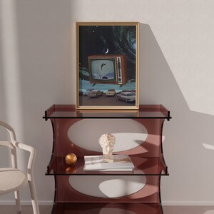 Puede incluir: Una impresi&oacute;n enmarcada de un televisor vintage con un dise&ntilde;o retro, que muestra una escena de coches conduciendo por una carretera con una luna creciente en el cielo. La impresi&oacute;n se muestra en una estanter&iacute;a de vidrio de tres niveles con un dise&ntilde;o redondo y ovalado.