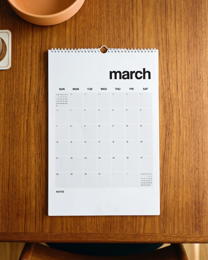 May include: A white wall calendar with black text showing the month of March. The calendar features a grid layout with days of the week labeled from Sunday to Saturday, a spiral binding at the top, and a dedicated section for notes at the bottom.