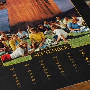 Pu&ograve; includere: Una pagina del calendario di settembre con una fotografia vintage di un gruppo di persone sedute su una collina erbosa vicino a una grande formazione rocciosa. La griglia del calendario &egrave; stampata in giallo su uno sfondo nero.