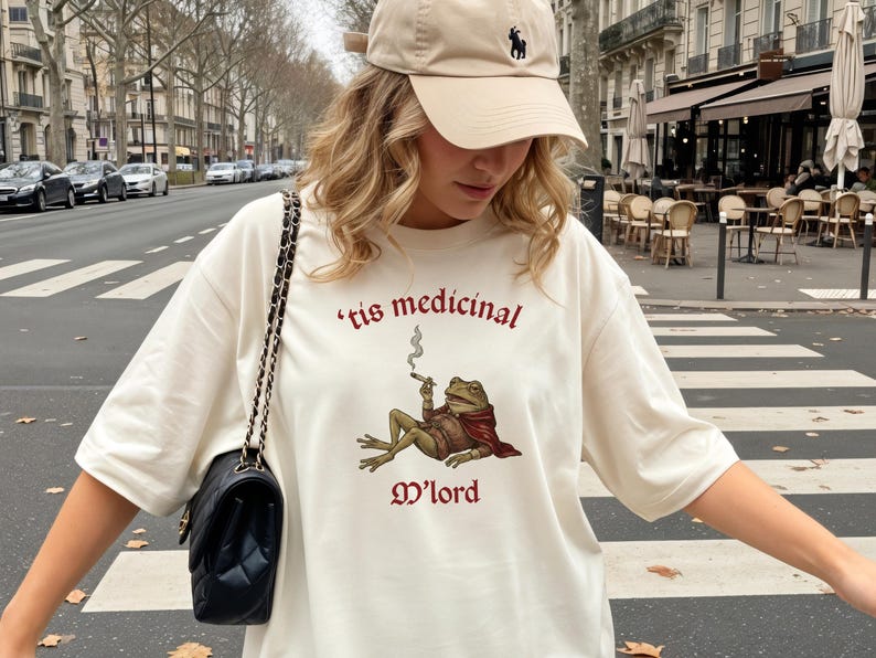 Może przedstawiać: Kremowa koszulka oversize z ilustracją żaby palącej papierosa, z bordowym napisem "'tis medicinal M'lord". Str&oacute;j uzupełnia beżowa czapka z daszkiem i czarna pikowana torba na ramię.