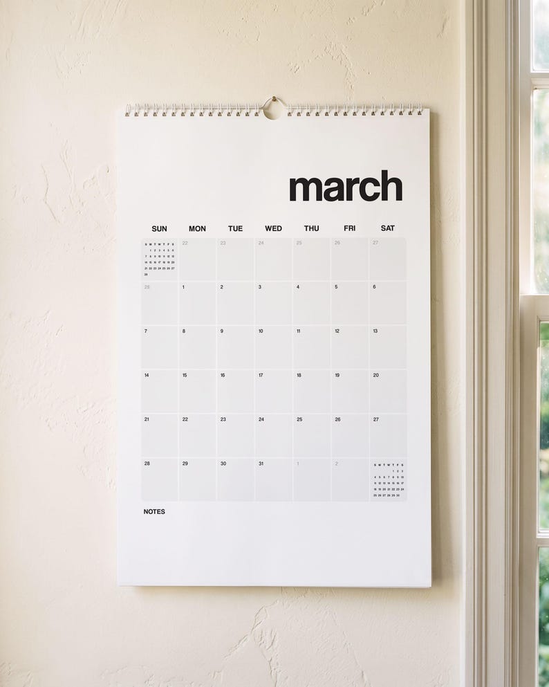 May include: A white wall-mounted calendar for the month of March with black sans-serif text. The calendar features a grid layout with days of the week labeled from Sunday to Saturday, a small reference calendar, and a designated section for notes.