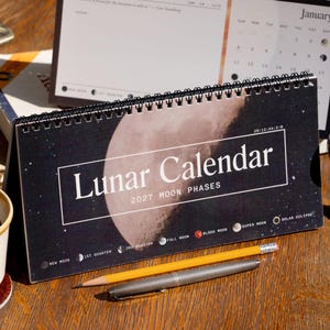 Può includere: Un calendario da scrivania 2027 Lunar Calendar con rilegatura a spirale nera, caratterizzato da una grande grafica lunare, il testo Lunar Calendar 2027 Moon Phases e una legenda delle fasi lunari. Una matita gialla e una penna in metallo argento.
