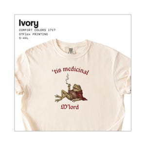 Może przedstawiać: Kremowa koszulka z grafiką żaby palącej papierosa, w pelerynie i napisem "'tis medicinal, M'lord". Koszulka to nadruk DTFlex Comfort Colors 1717, dostępny w rozmiarach S-4XL.