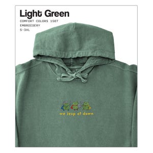 Może przedstawiać: Jasnozielona bluza z kapturem ze sznurkiem i haftowanym wzorem żaby. Pod spodem wyhaftowano napis "we leap at dawn". Bluza z kapturem jest oznaczona jako "Light Green" i jest dostępna w rozmiarach S-3XL.