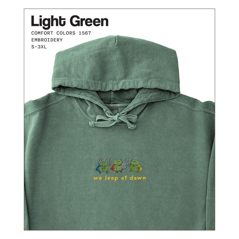 Może przedstawiać: Jasnozielona bluza z kapturem ze sznurkiem i haftowanym wzorem żaby. Pod spodem wyhaftowano napis "we leap at dawn". Bluza z kapturem jest oznaczona jako "Light Green" i jest dostępna w rozmiarach S-3XL.