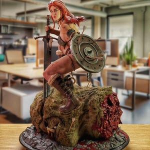 Könnte beinhalten: Eine rothaarige weibliche Kriegerfigur steht auf einem schädelförmigen Sockel. Sie hält ein Schwert und einen Schild und ist mit Blut und Pfeilen bedeckt. Die Figur ist aus dunkelgrauem Material gefertigt und etwa 30 cm hoch.