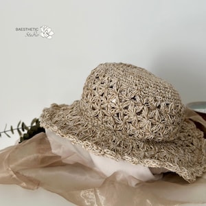 May include: A beige crochet sun hat with a wide brim. The hat features an openwork floral pattern and a scalloped edge. The hat is made of a natural fiber and is suitable for sun protection. The hat is displayed on a white surface.
