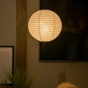 May include: A round, paper lantern hangs from the ceiling. The lantern is a warm, off-white color and has horizontal lines. The light inside the lantern glows, creating a soft, ambient light. The lantern is a decorative lighting fixture.