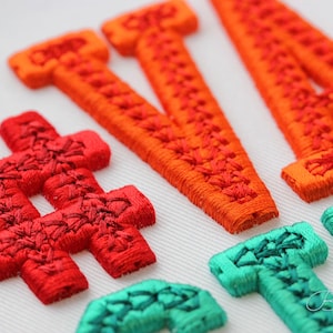 以下が含まれることがあります： 鮮やかな色の刺繍文字と記号のクローズアップ。文字はオレンジ色で、記号は赤とティールです。刺繍は白い生地に施されており、詳細なステッチが施されています。