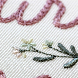 May include: Close-up of a delicate embroidered floral design with pink and green thread on a white fabric. The design features small flowers and leaves.