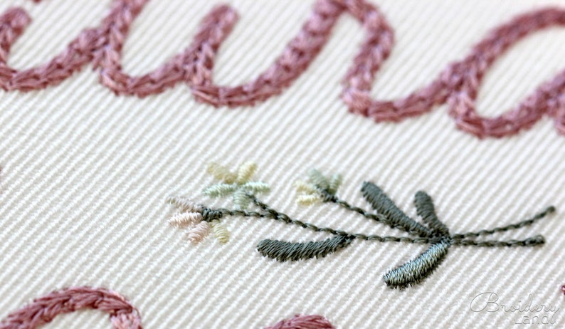 May include: Close-up of a delicate embroidered floral design with pink and green thread on a white fabric. The design features small flowers and leaves.