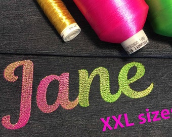 May include: The name "Jane" is embroidered in gold, pink, and green thread on a dark blue denim fabric. The word "XXL sizes!" is written in pink below the name. Three spools of thread are visible in the background: gold, pink, and green.