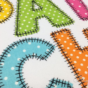 以下が含まれることがあります： カラフルな生地の文字のクローズアップ。それぞれがユニークな水玉模様で単語を綴っています。文字は白い背景に黒いステッチで縁取られています。文字は黄色、緑、ピンク、青、オレンジです。