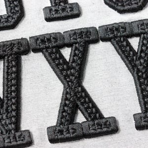 May include: Close-up of black embroidered letters on a white fabric background. The letters appear to be part of a larger word or phrase, with the letters W, X, and Y prominently displayed. The embroidery has a textured appearance.