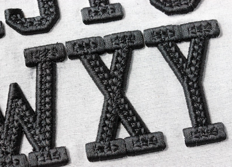 May include: Close-up of black embroidered letters on a white fabric background. The letters appear to be part of a larger word or phrase, with the letters W, X, and Y prominently displayed. The embroidery has a textured appearance.