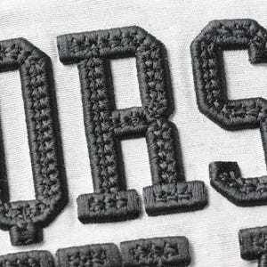 May include: Close-up of black embroidered letters on a white fabric background. The letters appear to be part of a larger word or phrase, with a textured, stitched appearance. The letters are in a bold, block style.