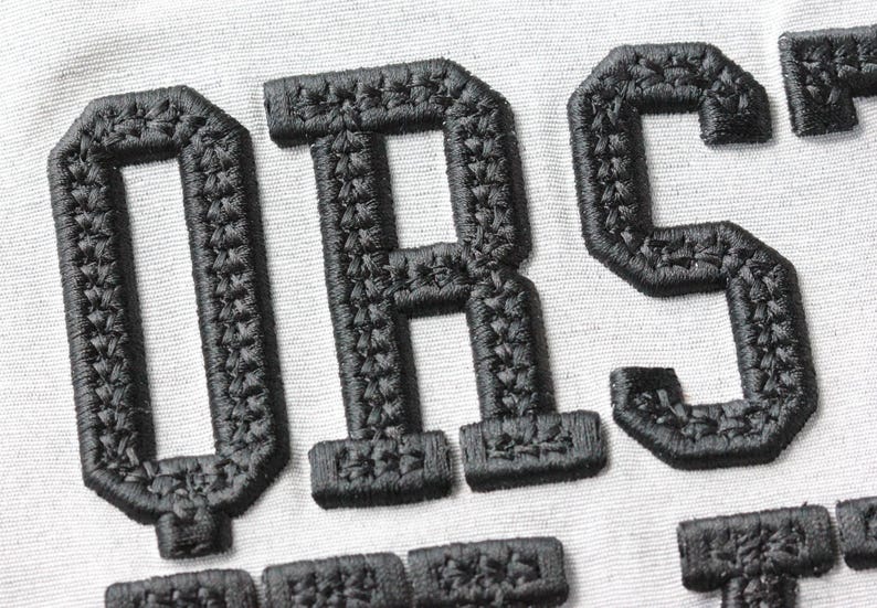 May include: Close-up of black embroidered letters on a white fabric background. The letters appear to be part of a larger word or phrase, with a textured, stitched appearance. The letters are in a bold, block style.