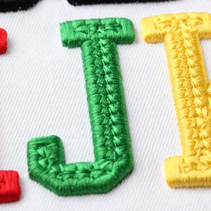 May include: Close-up of embroidered letters I, J, and K in red, green, and yellow, respectively, on a white fabric background. The letters are stitched with a textured thread, creating a raised effect. The image highlights the detailed embroidery work.