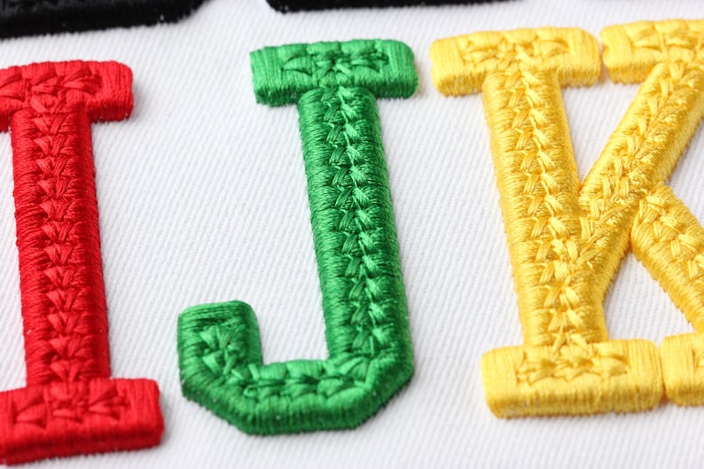 May include: Close-up of embroidered letters I, J, and K in red, green, and yellow, respectively, on a white fabric background. The letters are stitched with a textured thread, creating a raised effect. The image highlights the detailed embroidery work.