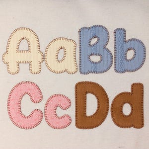 Könnte beinhalten: Ein Set aus Stoffbuchstaben mit den Groß- und Kleinbuchstaben A, B, C und D. Die Buchstaben sind mit sichtbaren braunen Nähten an den Rändern appliziert. Das A ist cremefarben, das B ist blau mit einer feinen Textur, das C ist rosa und das D ist braun.